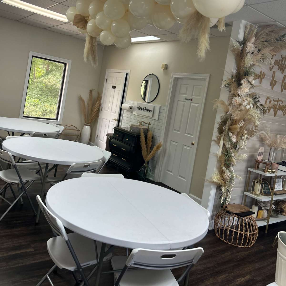 A decorated indoor event space with white tables and a balloon arrangement overhead, complemented by dried floral accents on the wall.