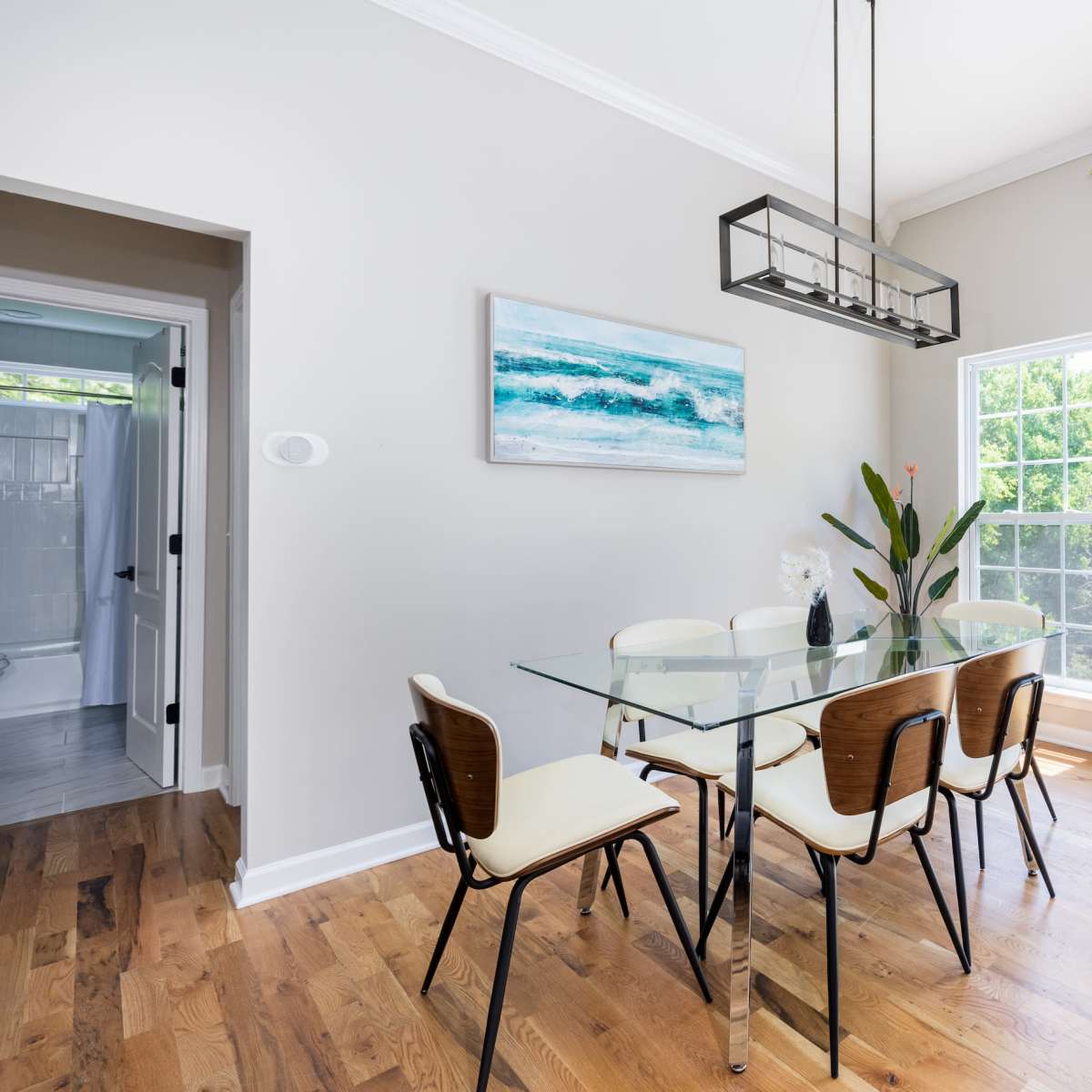 A modern dining area features a glass table with six chairs, a framed ocean painting on the wall, and large windows providing natural light and a view of greenery.