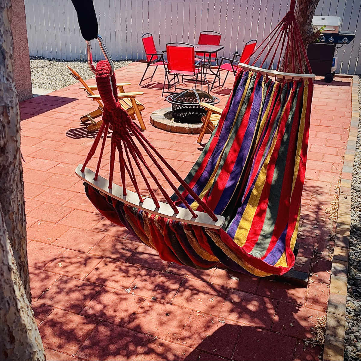 A colorful hammock hangs between two trees in a patio area with red chairs and a fire pit.