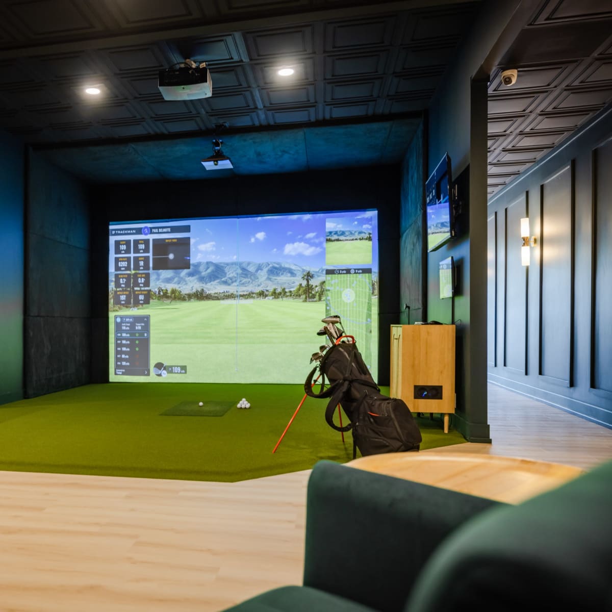 An indoor golf simulation room with a golf simulator screen, green turf, and golf equipment, surrounded by a seating area and a table.