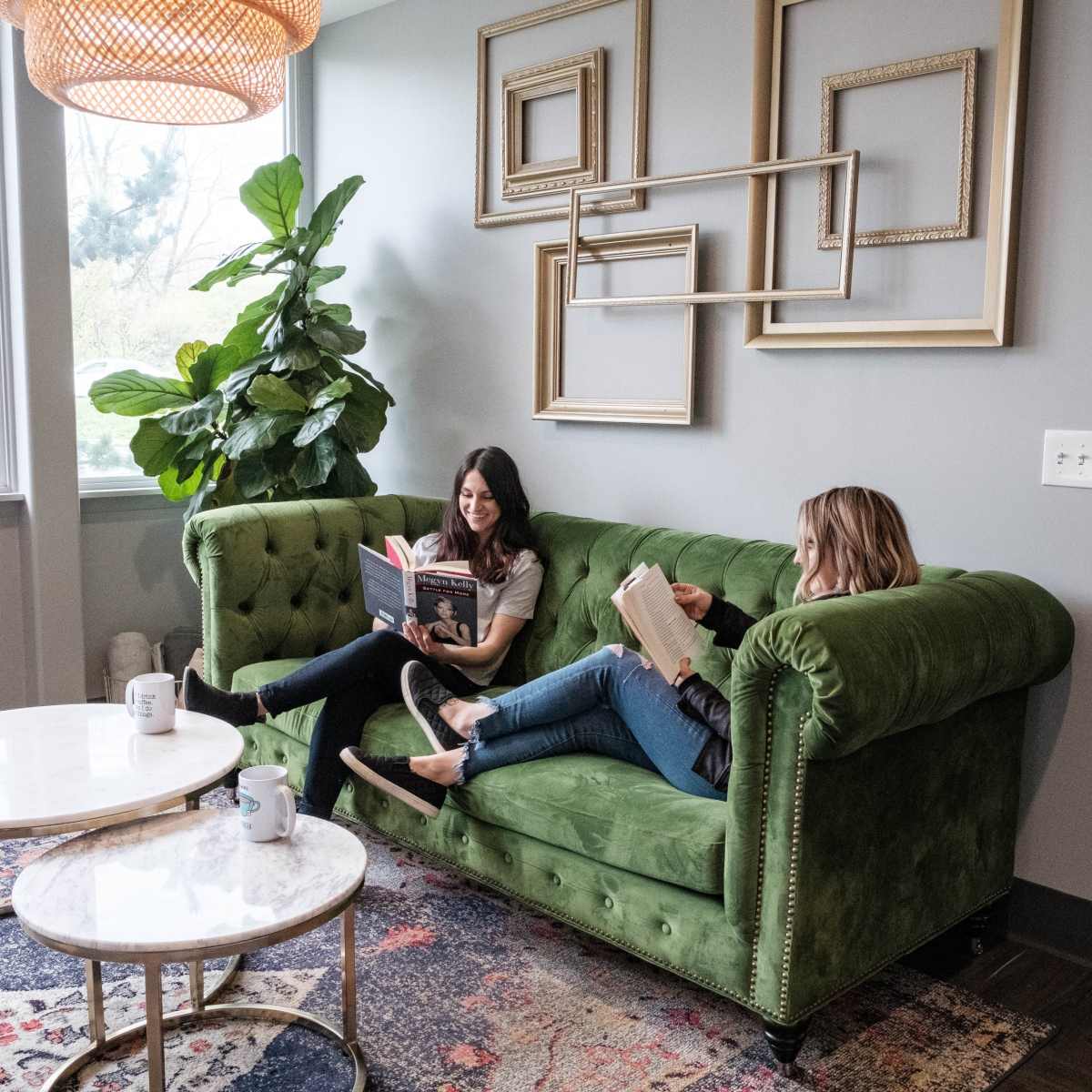 Two women are sitting on a green couch in a bright room, reading books while surrounded by plants and framed art.