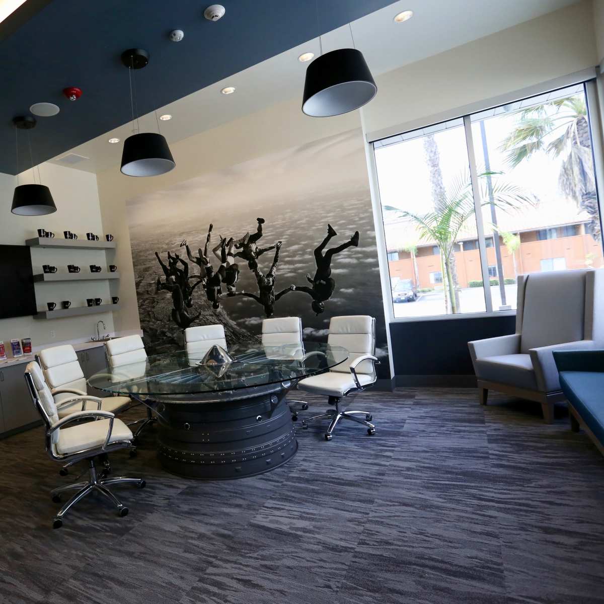 A modern conference room with a round glass table surrounded by white chairs, a wall decorated with a black-and-white mural, and a kitchenette area along one side.
