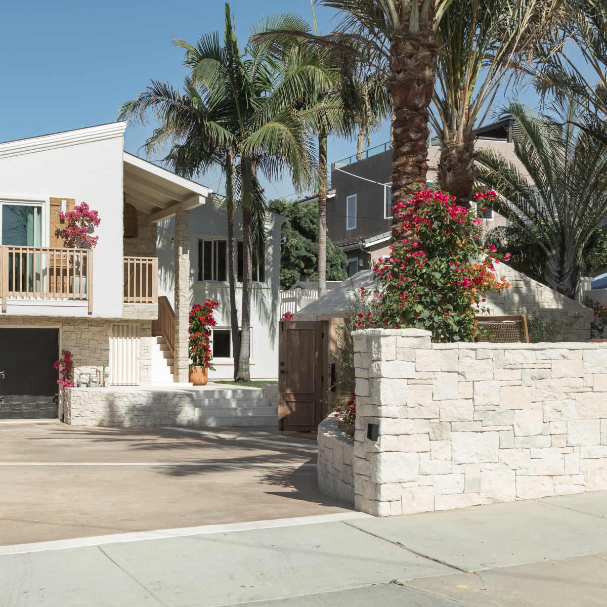 A modern two-story house with a front balcony is surrounded by palm trees and vibrant flowers in a residential area.