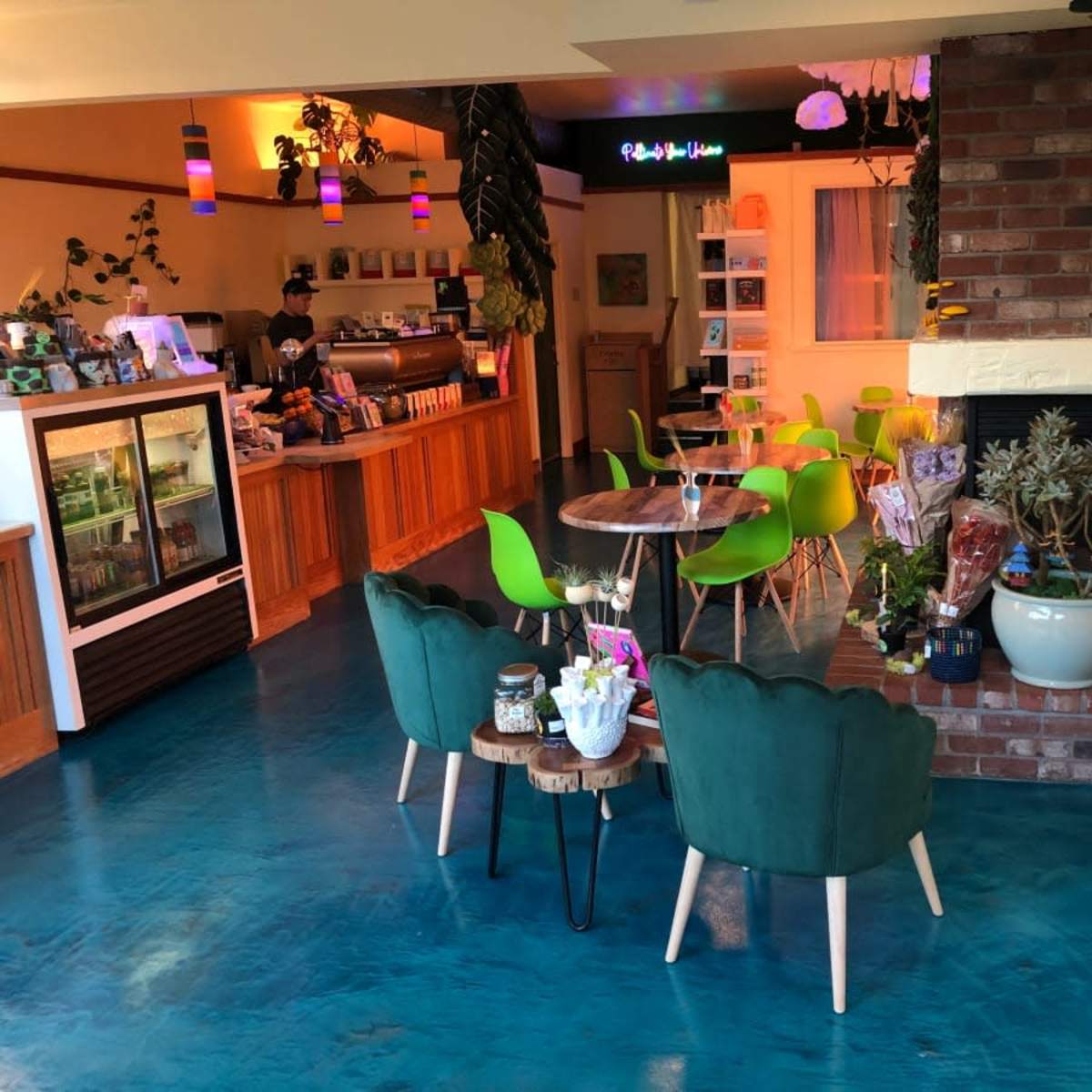 A modern cafe interior with green chairs, wooden tables, and a decorative wall featuring plants and artwork.