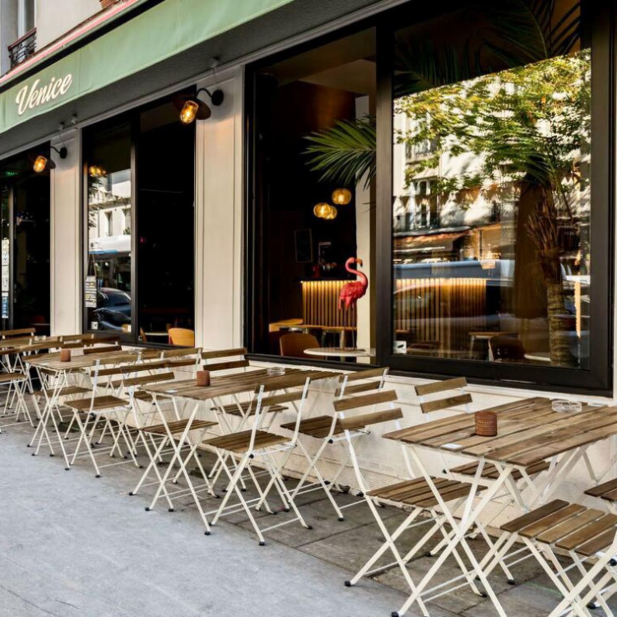 A restaurant patio with small wooden tables and chairs is situated on a sidewalk, next to large glass windows and a green awning.