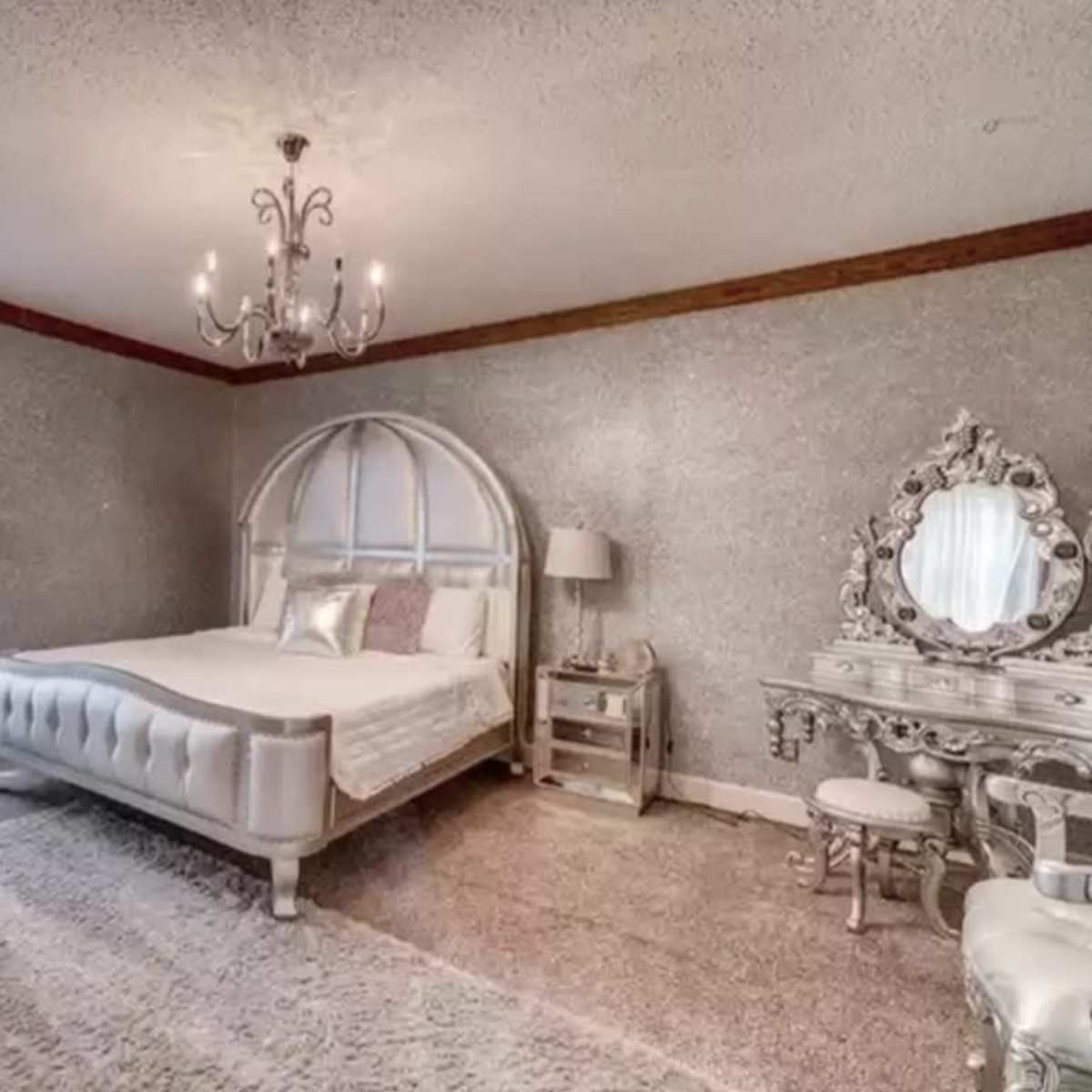 A spacious bedroom featuring a large bed, a decorative chandelier, a mirrored vanity, and a window with sheer white curtains.