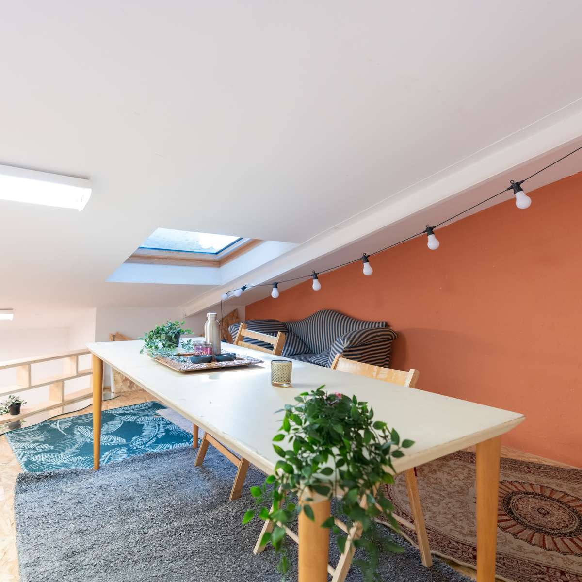 An attic space features a long table surrounded by chairs, with a cozy seating area and decorative plants under a skylight.