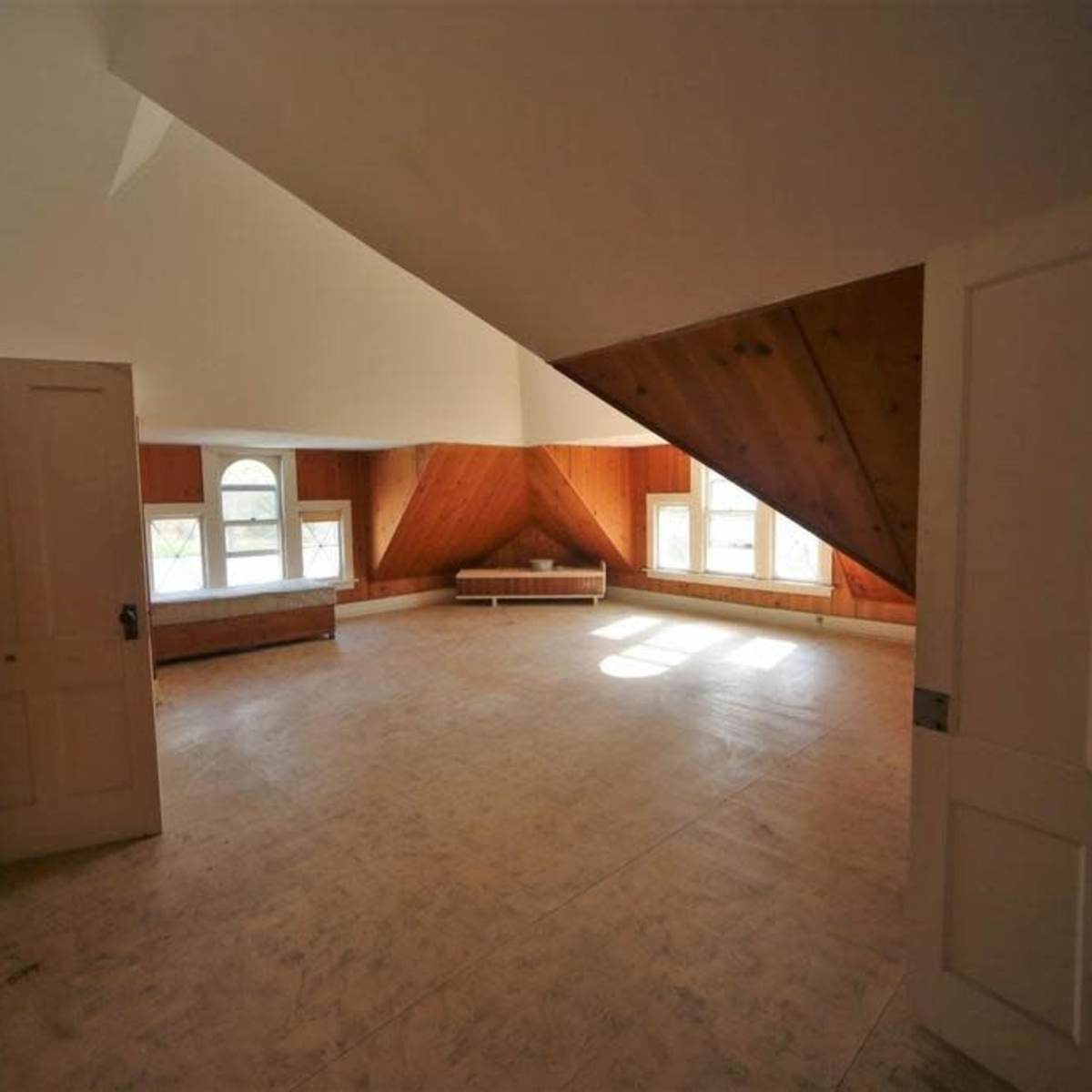 An empty attic room with slanted walls, wooden paneling, and two windows, illuminated by natural light.