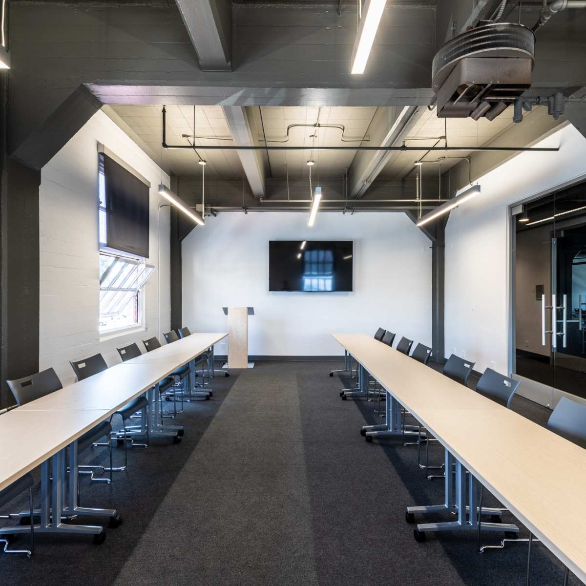A long, modern conference room features two rows of tables facing a screen, with chairs arranged on either side.