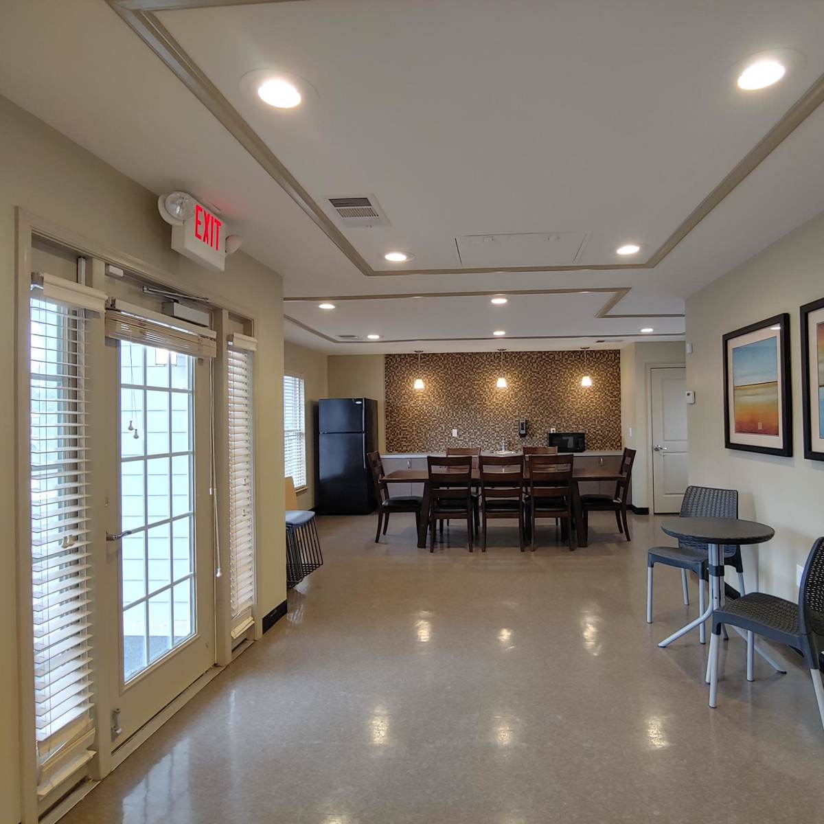 A modern communal kitchen area featuring a dining table, chairs, and a kitchenette with a refrigerator, as well as access to an outdoor patio through double doors.
