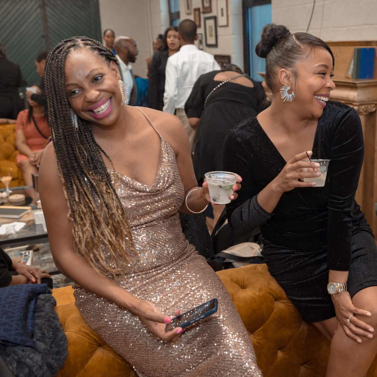 Two women sit on a velvet couch in a social gathering, each holding a drink and smiling while surrounded by other guests.