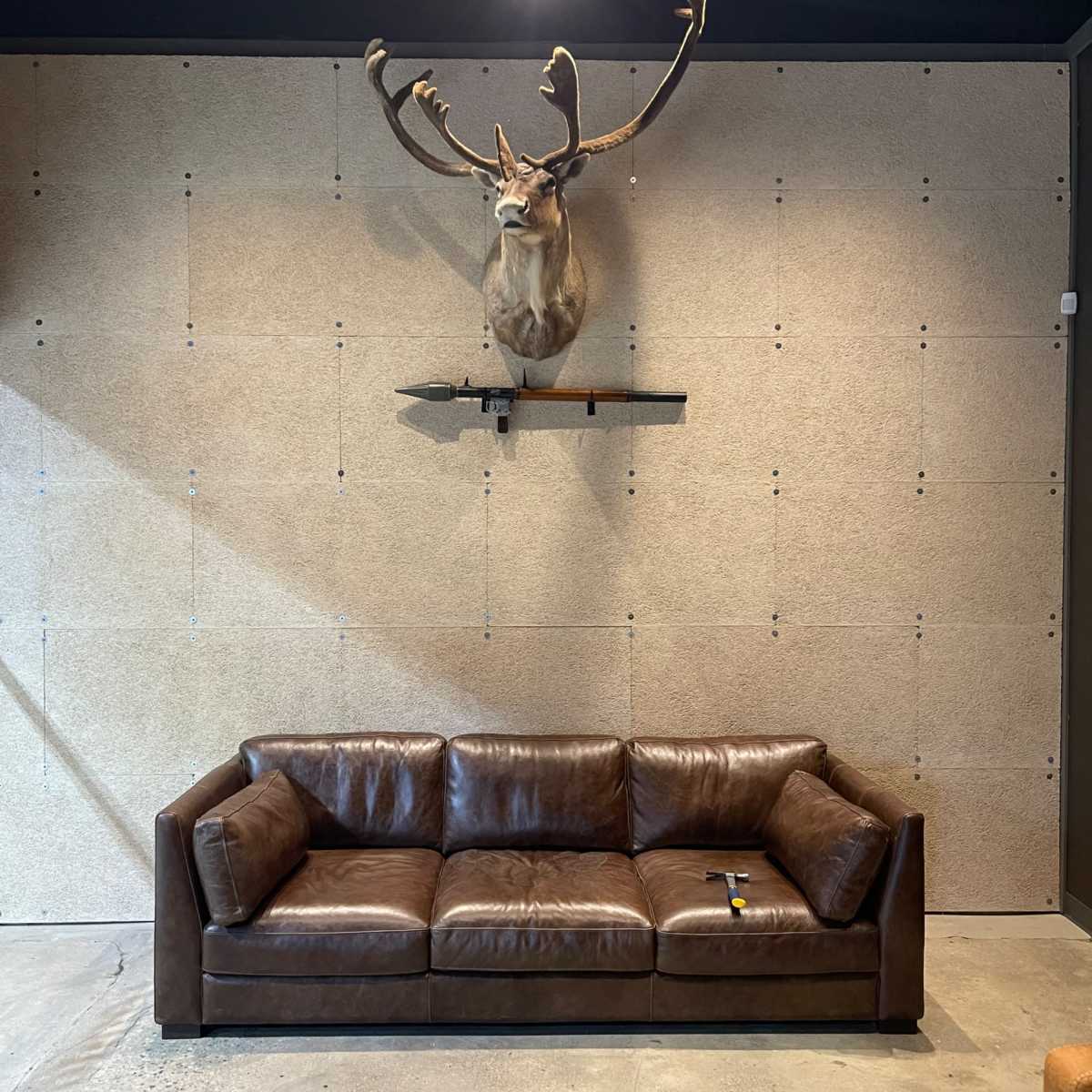 A brown leather sofa positioned against a textured wall, with a deer head mounted above it on a shelf.