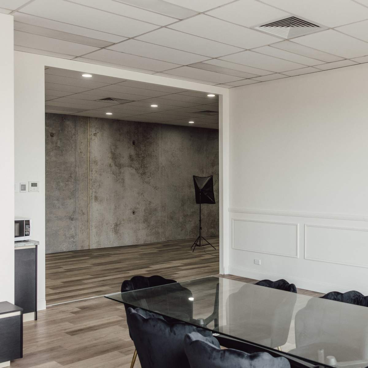 A modern office space features a glass dining table surrounded by black chairs, with an adjacent room showcasing a concrete wall and studio lighting setup.