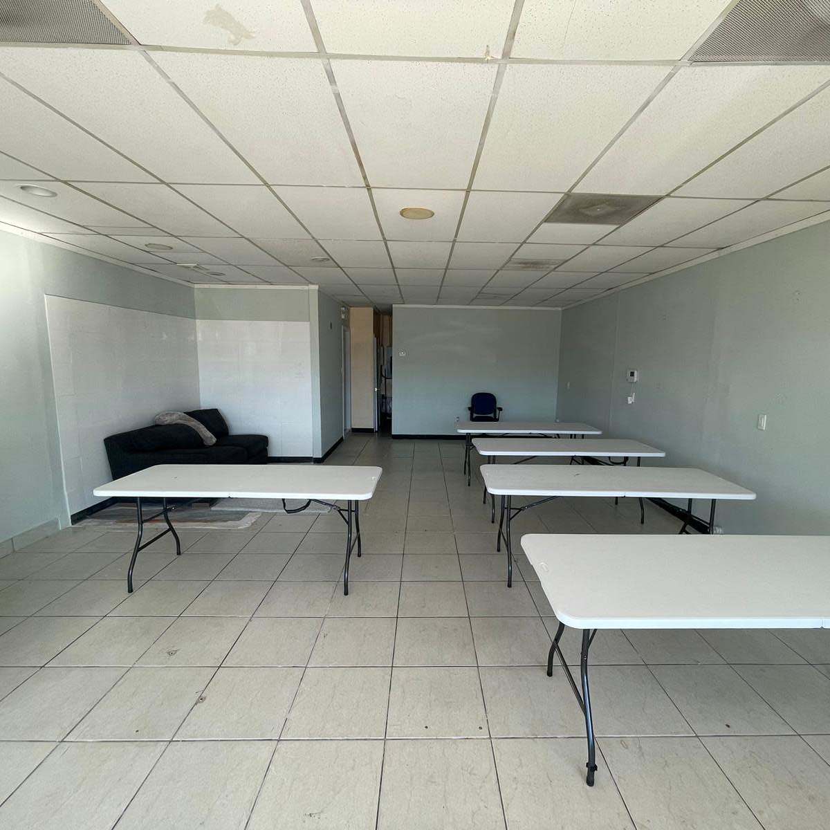 An empty room with several white folding tables arranged around a couch and a chair in the background.
