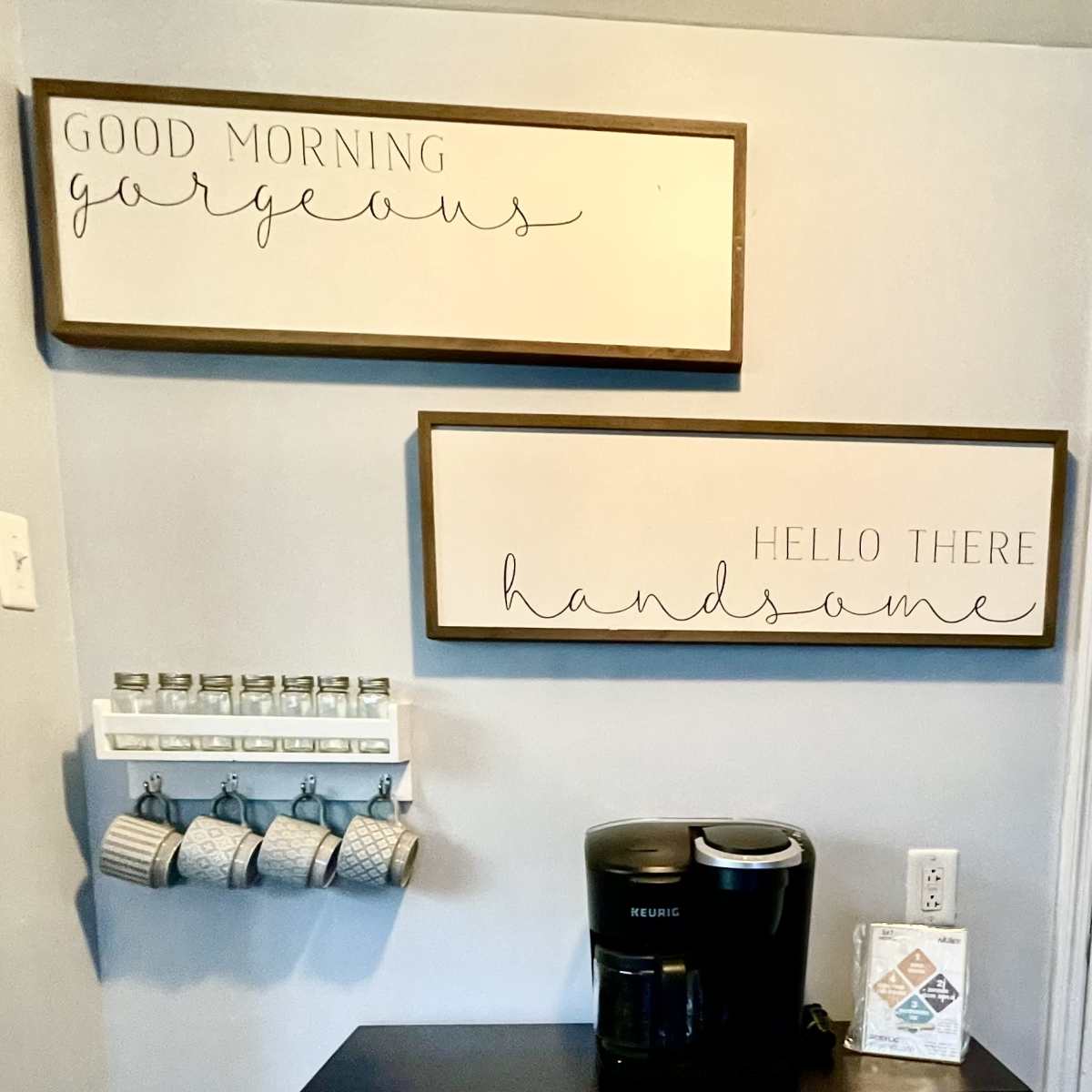 A coffee station with a Keurig machine, decorative signs on the wall, and a rack holding cups.