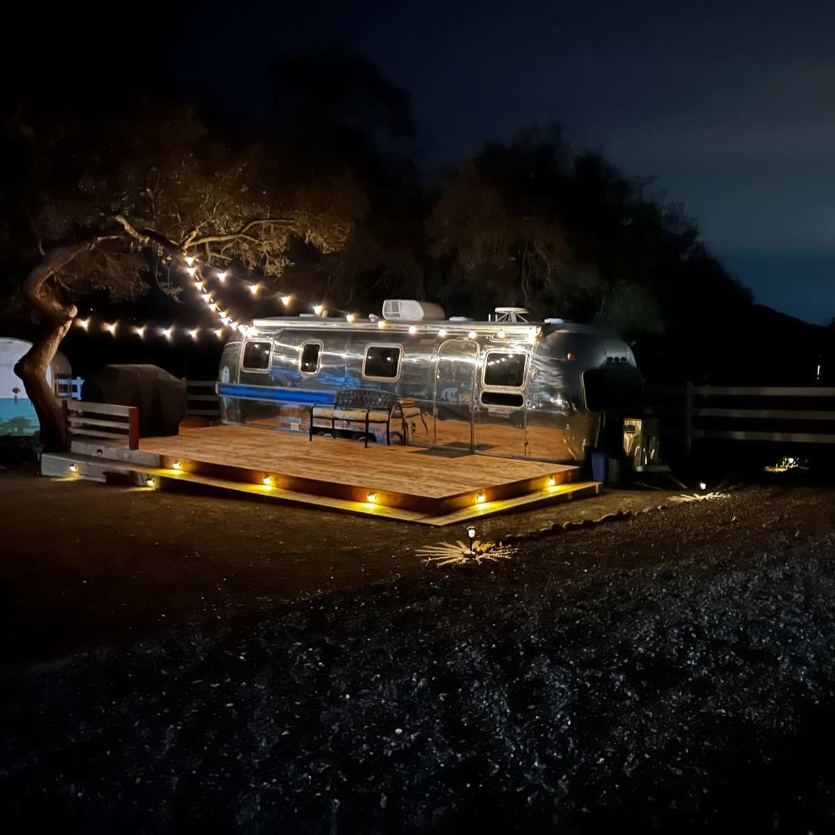 An Airstream trailer is parked on a wooden deck illuminated by string lights in a dark outdoor setting.