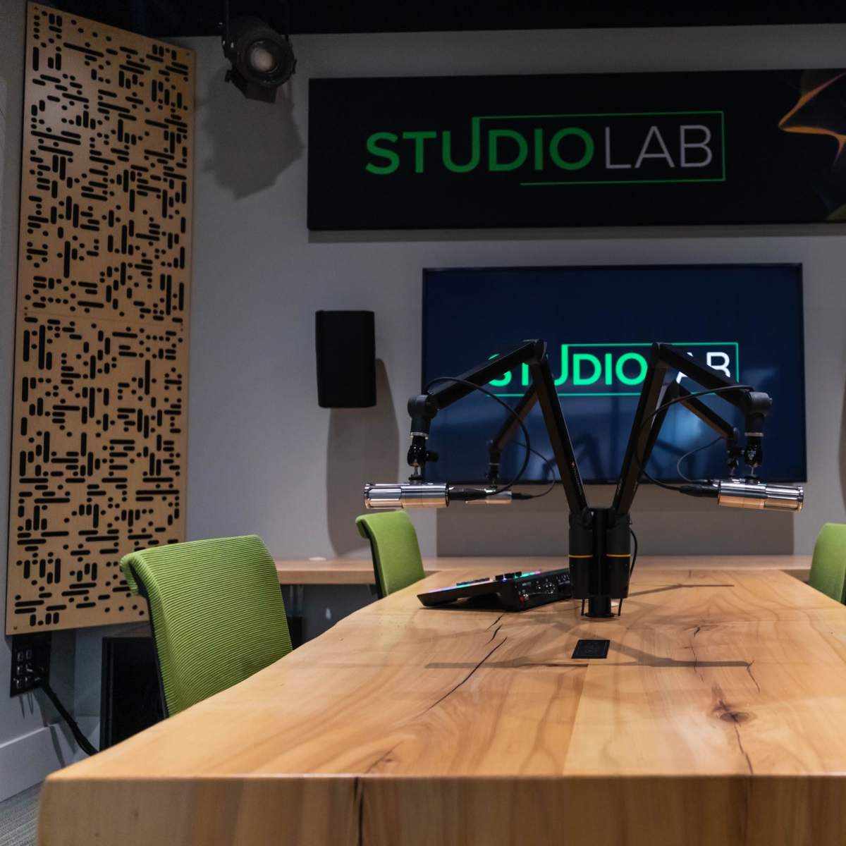 A modern podcast studio featuring a large wooden table, two green chairs, and a wall-mounted television with the "STUDIOLAB" logo.