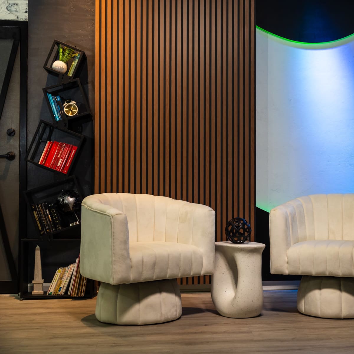 Two white upholstered chairs positioned across from a small white table in a modern interior space featuring a textured wall and colorful ambient lighting.