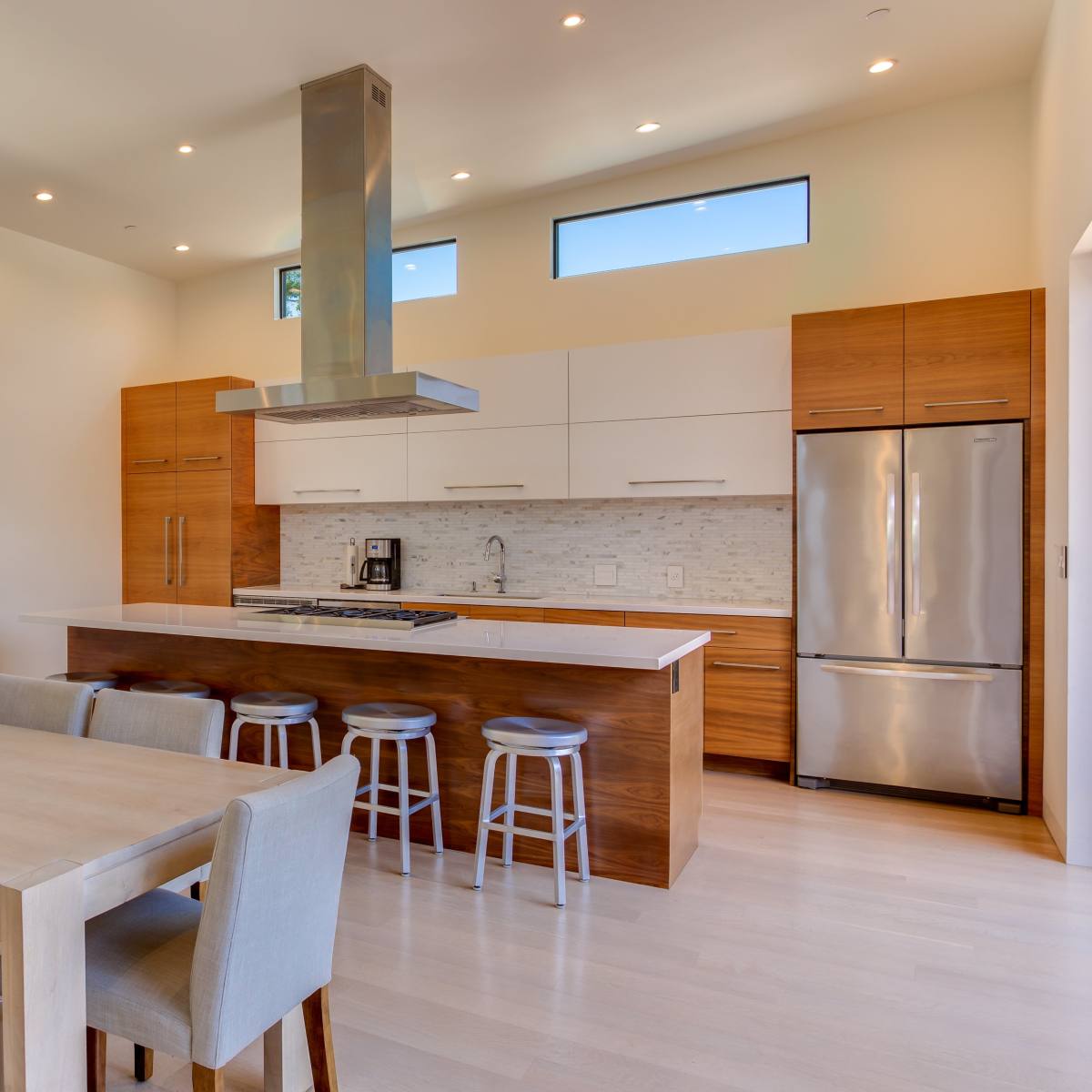 A modern kitchen and dining area featuring a central island with bar stools, a wooden dining table, and large windows for natural light.