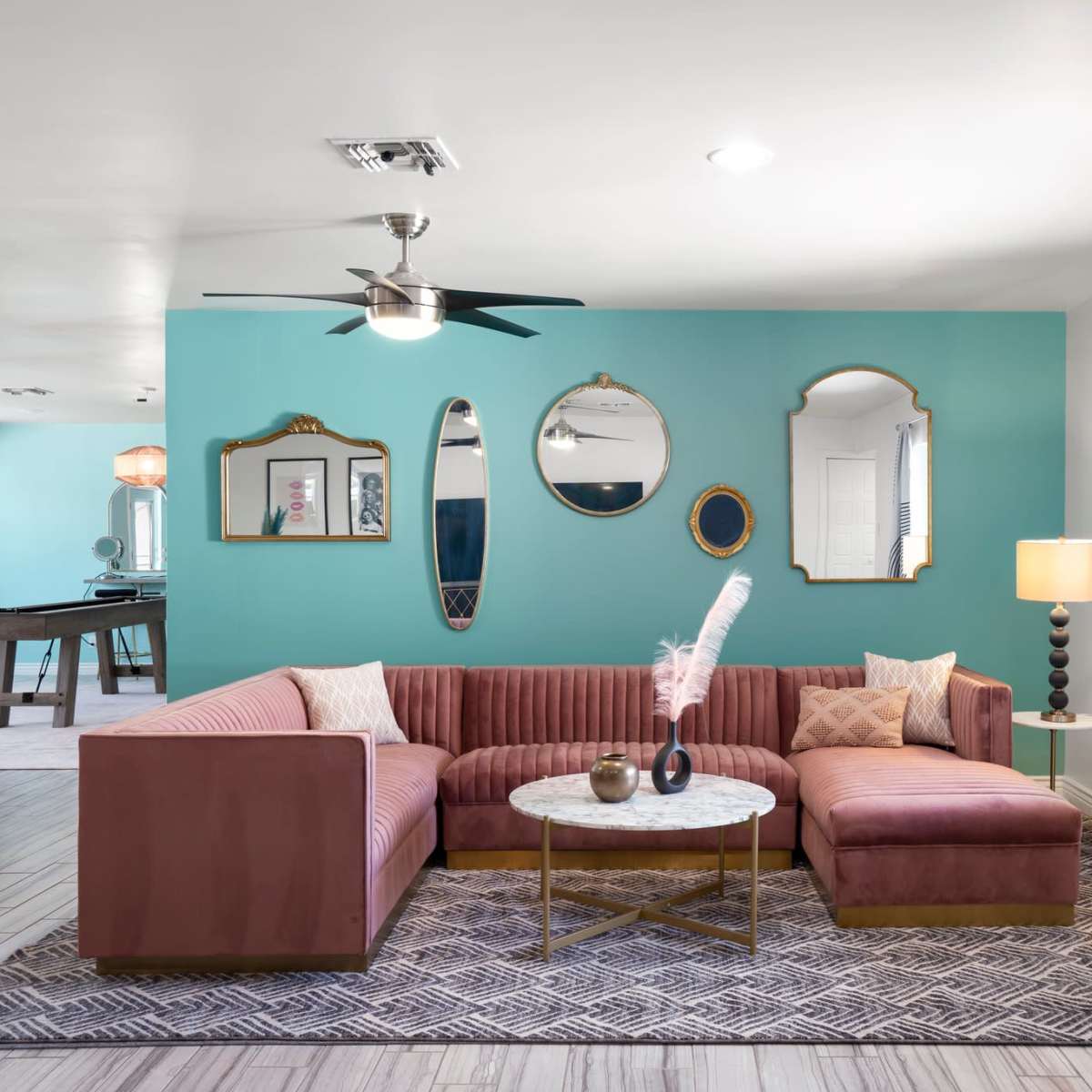 A modern living room with a large pink sectional couch, a round coffee table, and turquoise walls, featuring decorative mirrors and a lamp.