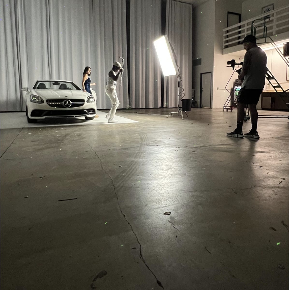 A man and a woman pose in front of a white Mercedes-Benz in a studio with draped curtains and a bright lighting setup.