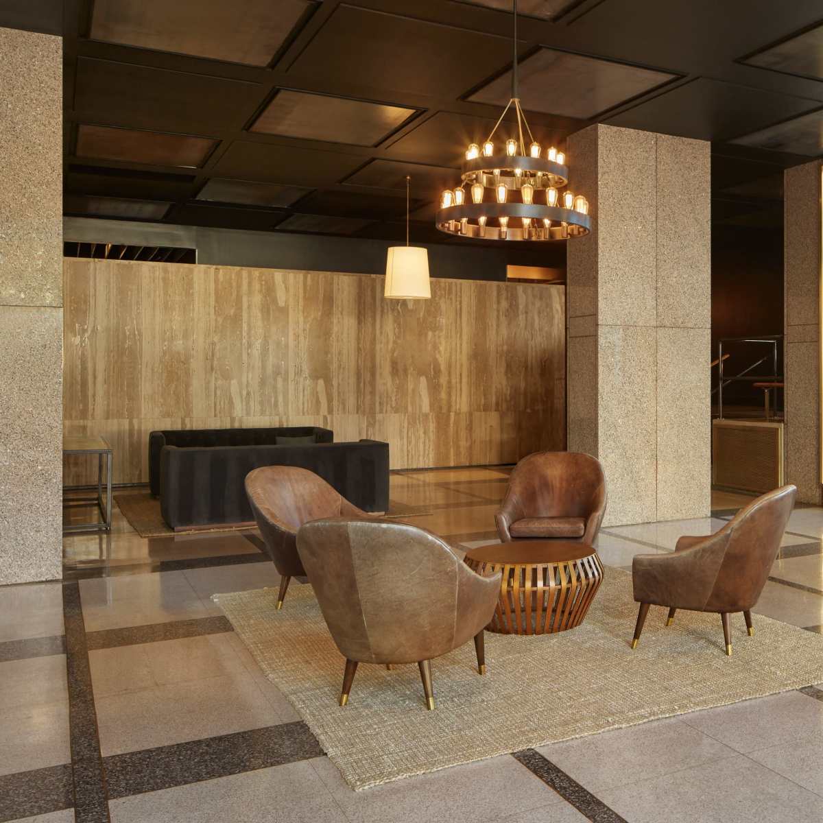 A modern lobby area featuring a seating arrangement of four brown armchairs around a circular wooden table on a light rug, with large windows allowing natural light to fill the space.