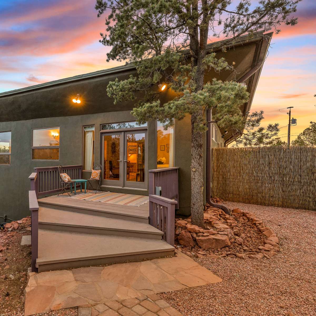 A modern house with large windows and a deck sits among trees at sunset, reflecting vibrant colors in the sky.