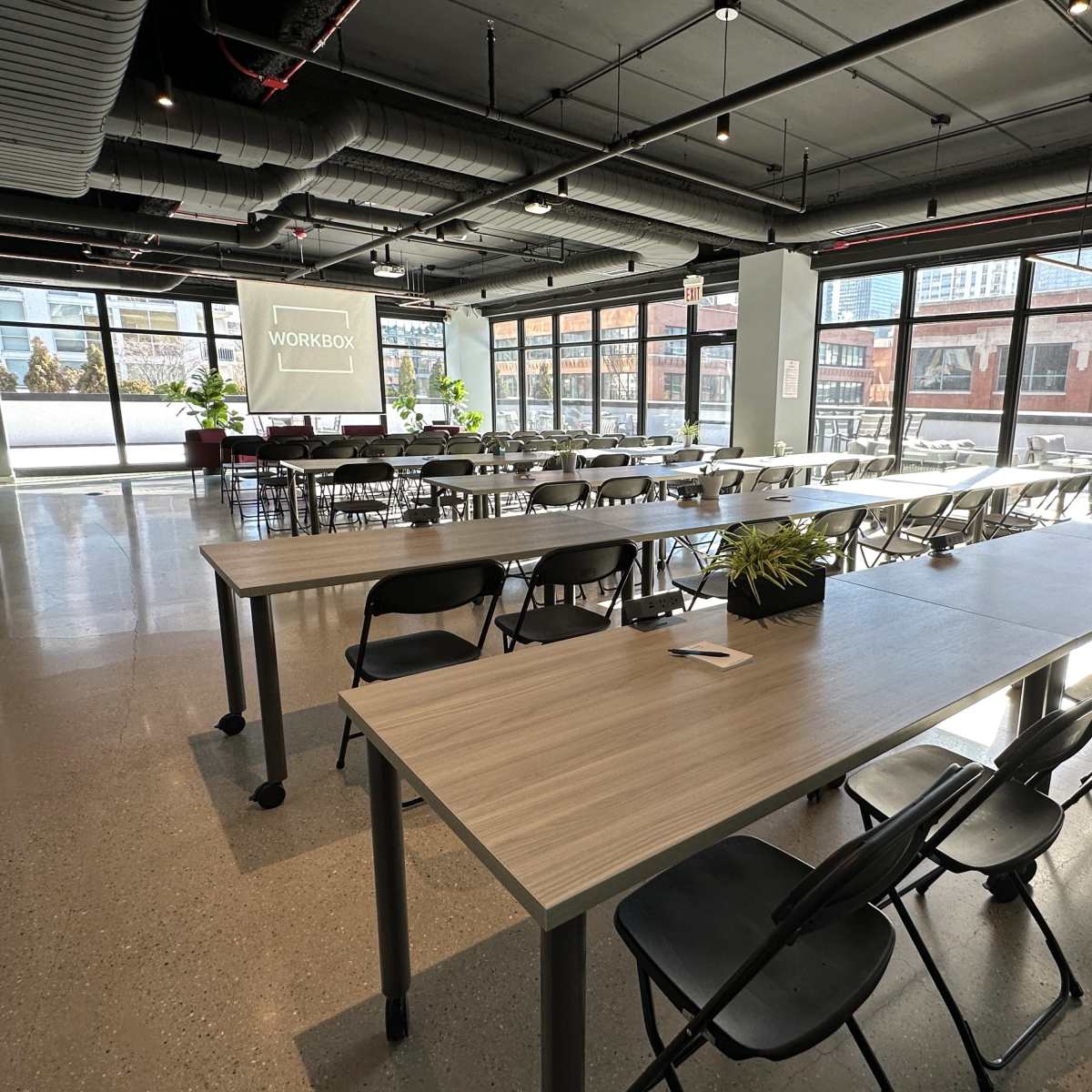 A spacious open office area with long tables and rows of chairs, featuring large windows that let in natural light.
