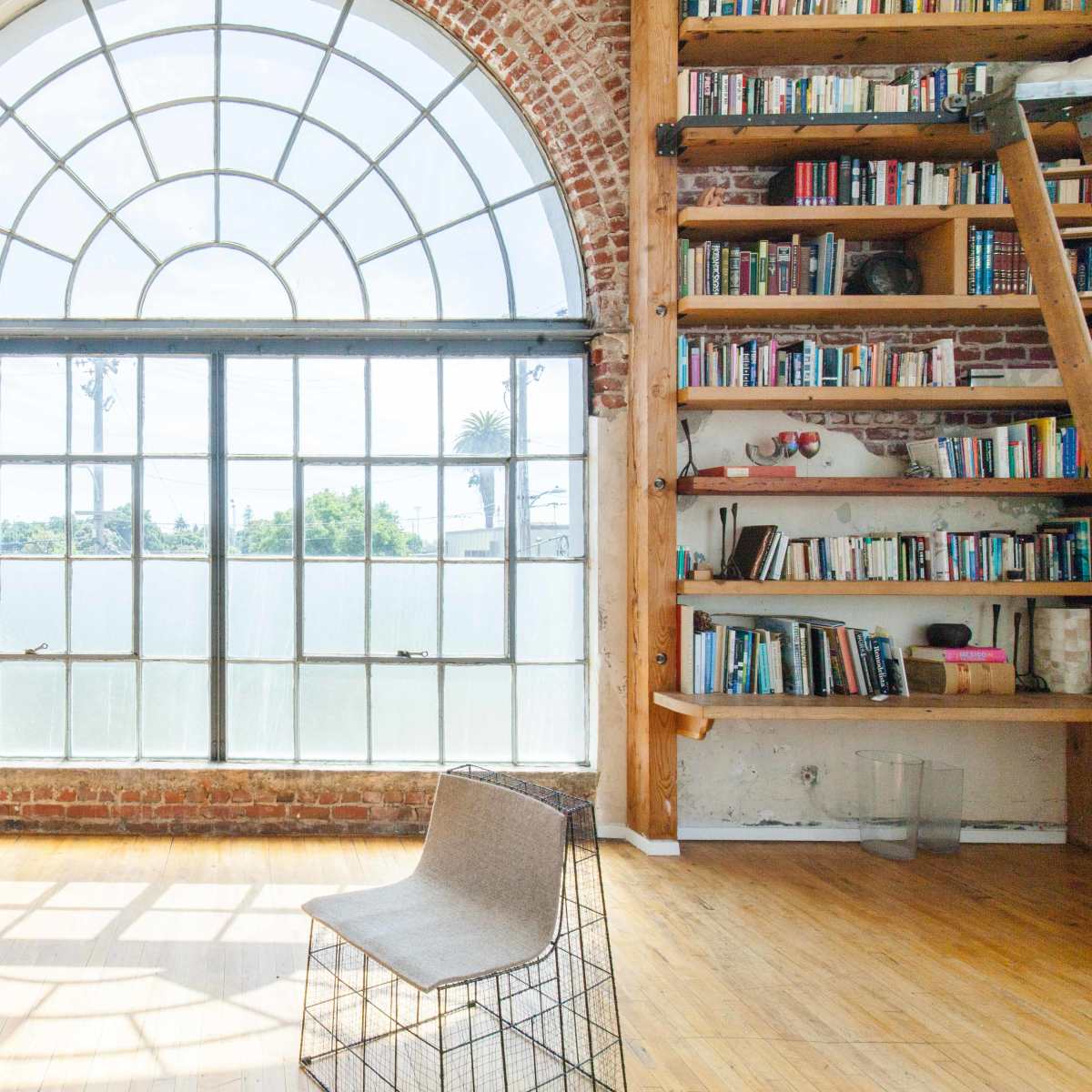 A spacious room features a large arched window, wooden flooring, and a bookshelf against a brick wall, with a single chair positioned near the window.