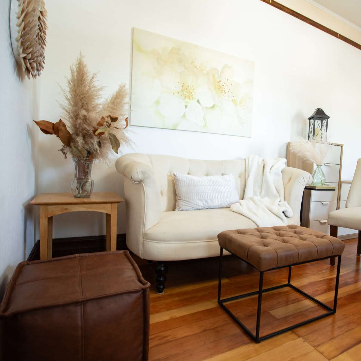 A cozy living room with a beige tufted sofa, a small wooden side table, a decorative armchair, and a brown leather ottoman, all situated on polished wooden floors.