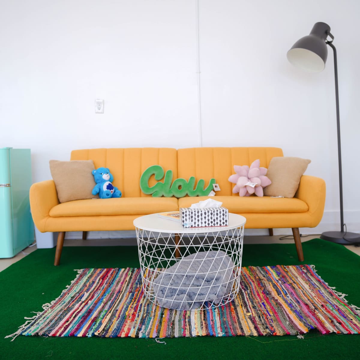 A bright yellow couch with decorative pillows sits on a green rug in a room featuring a small turquoise refrigerator and a round coffee table.