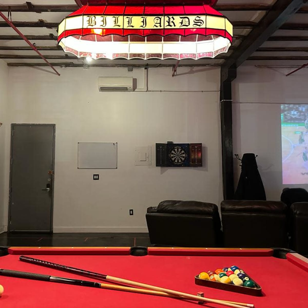 A pool table with colorful balls is illuminated by a hanging lamp in a game room featuring a dartboard and a basketball hoop.