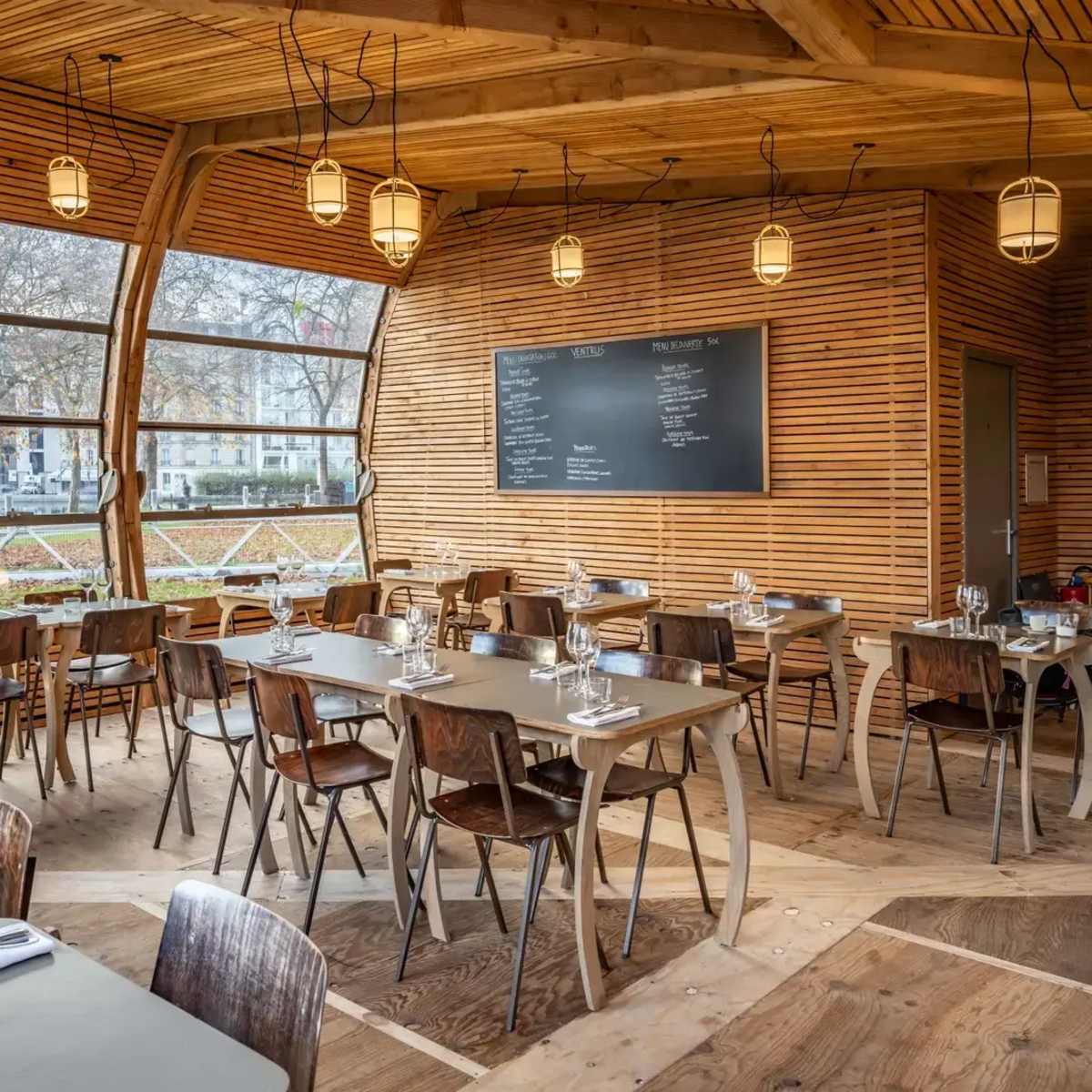 A cozy restaurant interior featuring wooden walls, tables set for dining, and large windows overlooking an outdoor view.