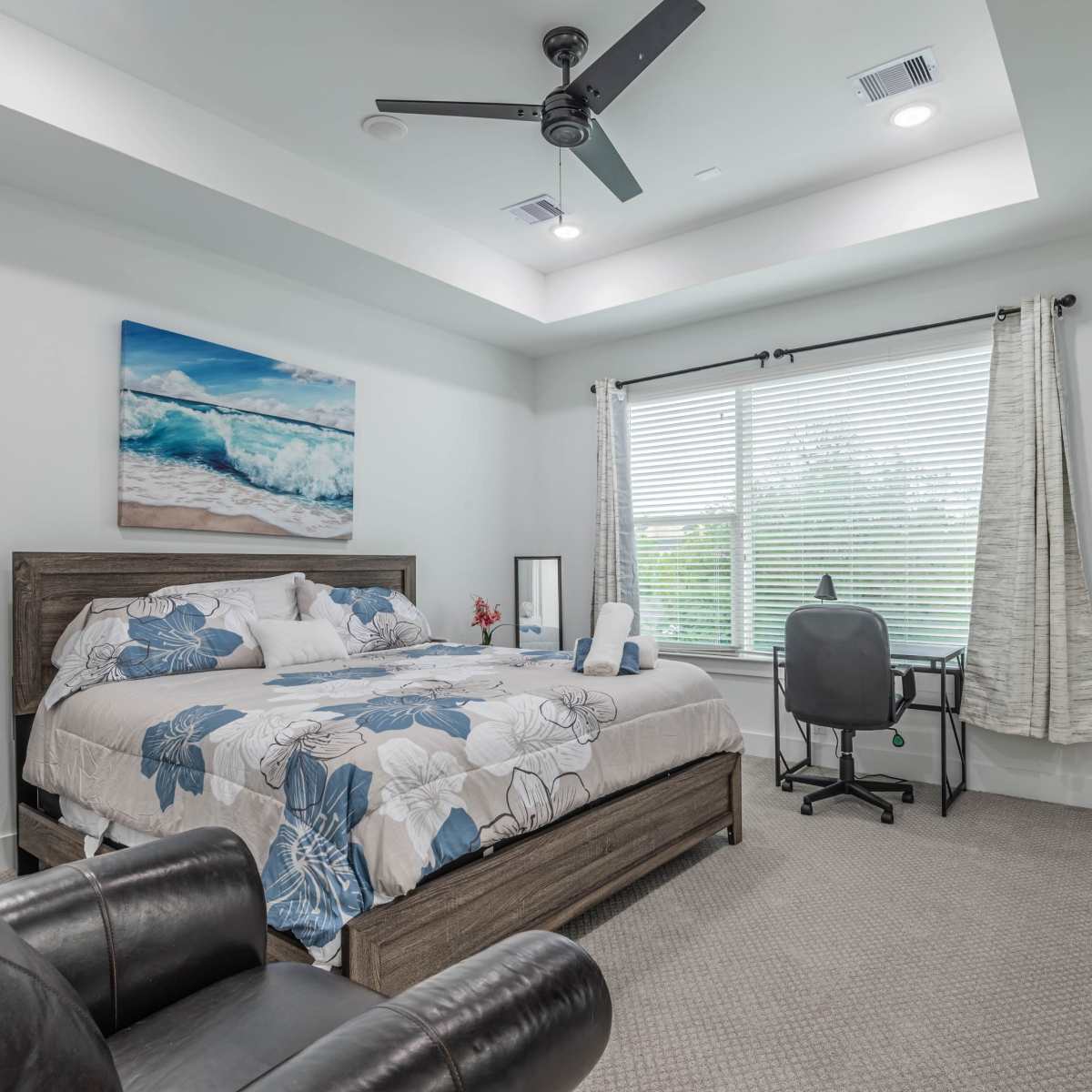 A modern bedroom with a large bed, desk, and a window dressed in light curtains, featuring a white ceiling and a ceiling fan.