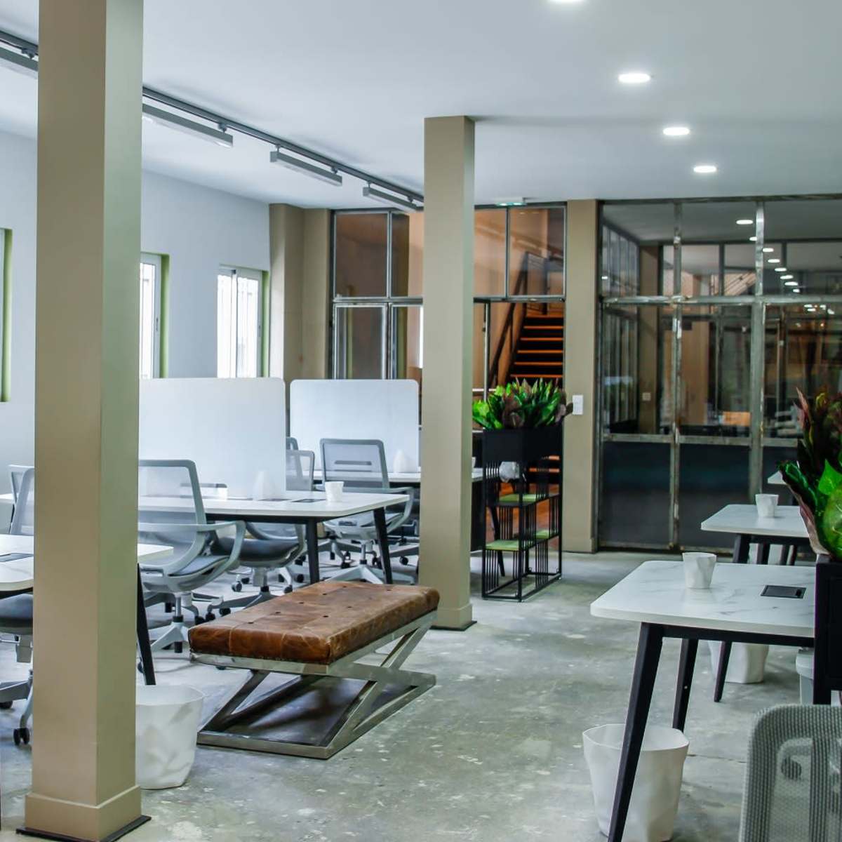 A modern office space with multiple workstations separated by dividers, large windows allowing natural light, and a mix of seating options.