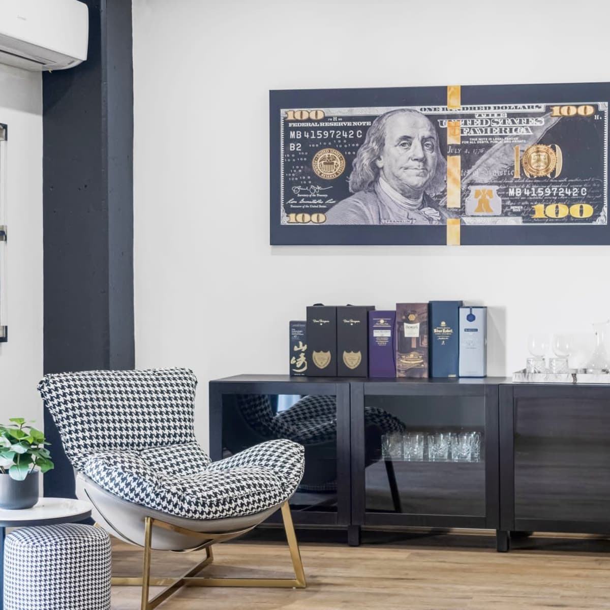 A modern office space featuring a black cabinet, a checkered armchair, a wall art piece of a $100 bill, and a small table with a plant.