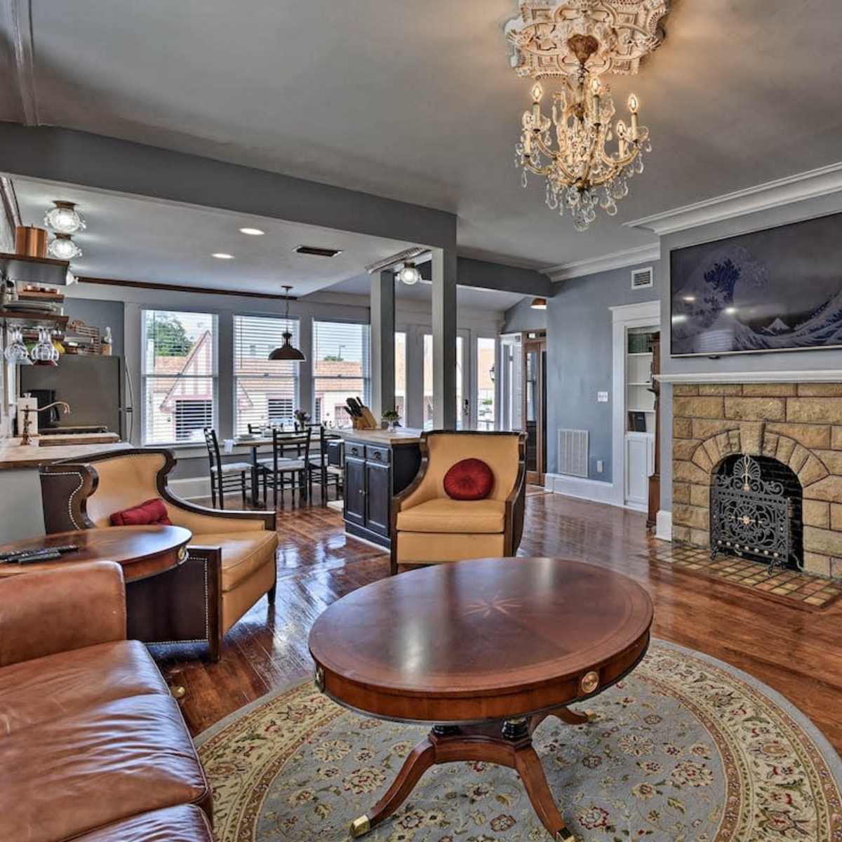 A living room featuring a chandelier, a circular rug, a vintage trunk, a stone fireplace, and large windows overlooking an outdoor view.