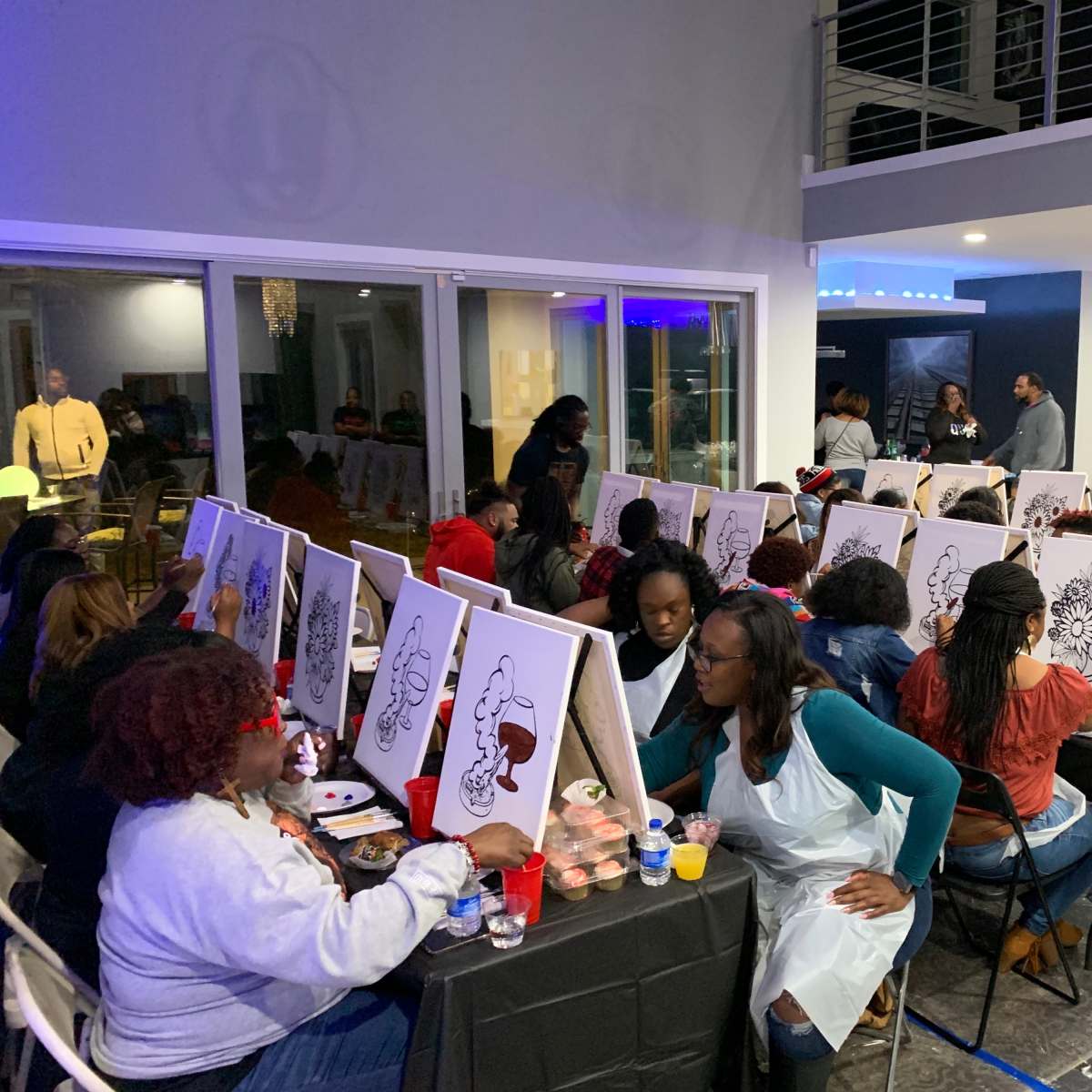 A group of people participate in a painting class, each working on canvas in a well-lit indoor space.