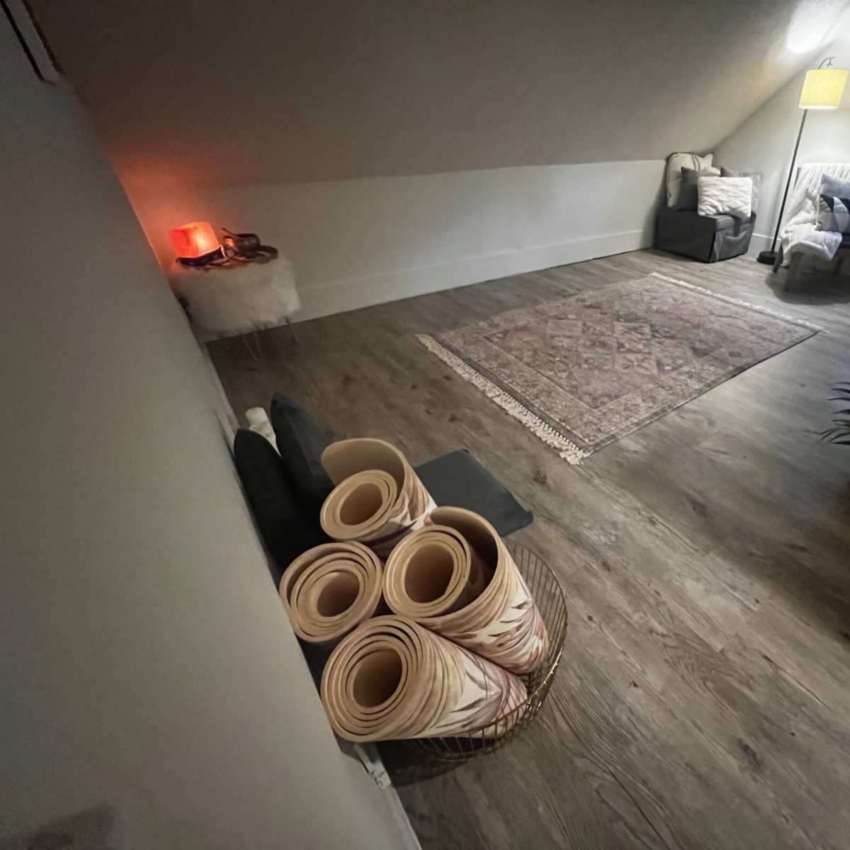A spacious, well-lit attic room features a woven rug, a small sofa, a chair, and neatly rolled mats in a basket.