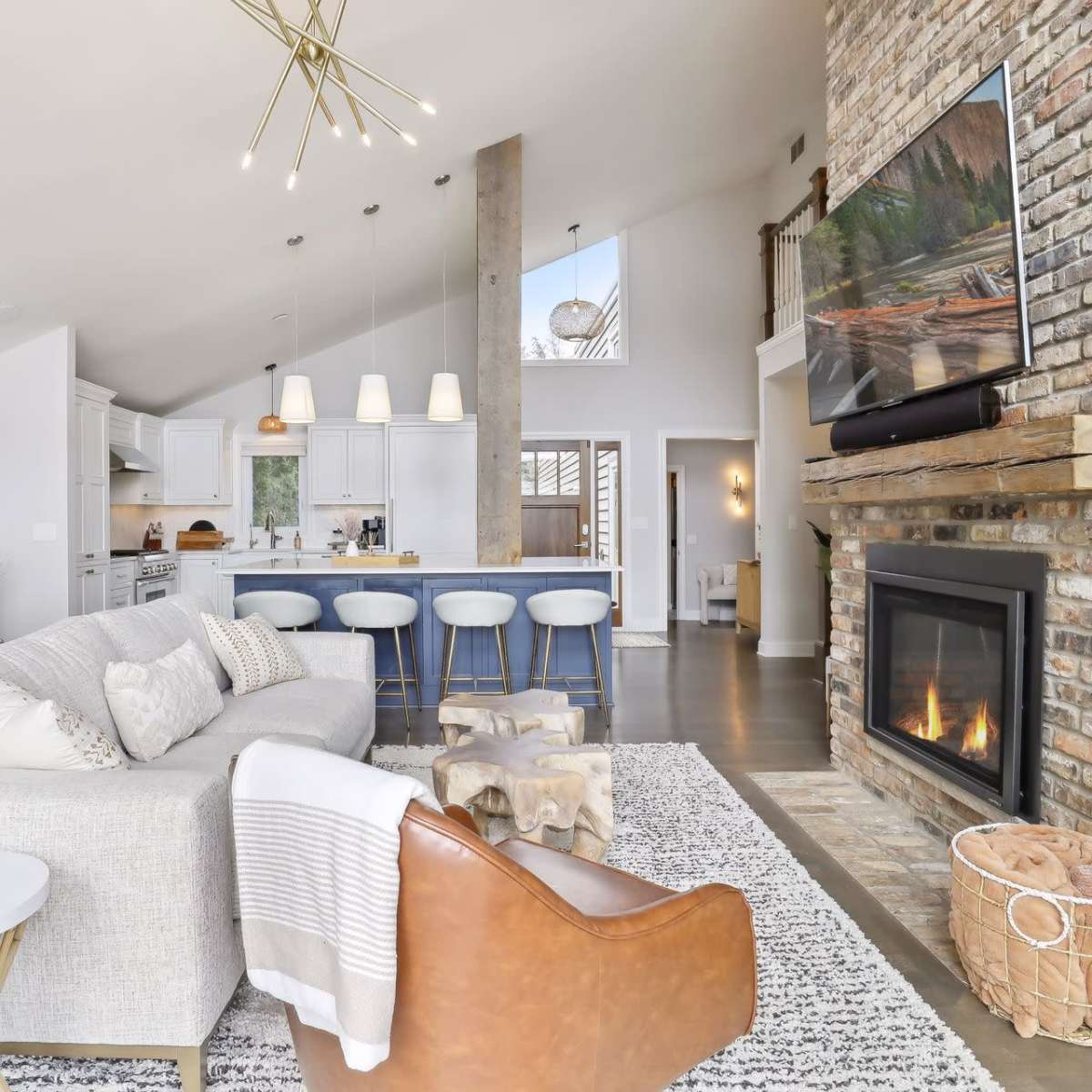 A modern living room features a cozy seating arrangement with a fireplace, a large TV mounted on a brick wall, and an open kitchen in the background.