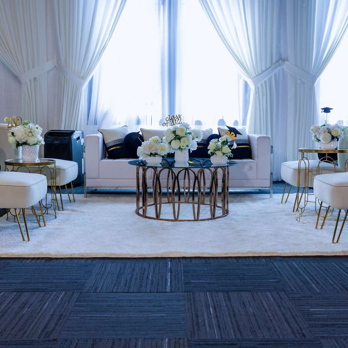 A stylish lounge area with white seating, a round coffee table, floral arrangements, and large windows covered by sheer curtains.
