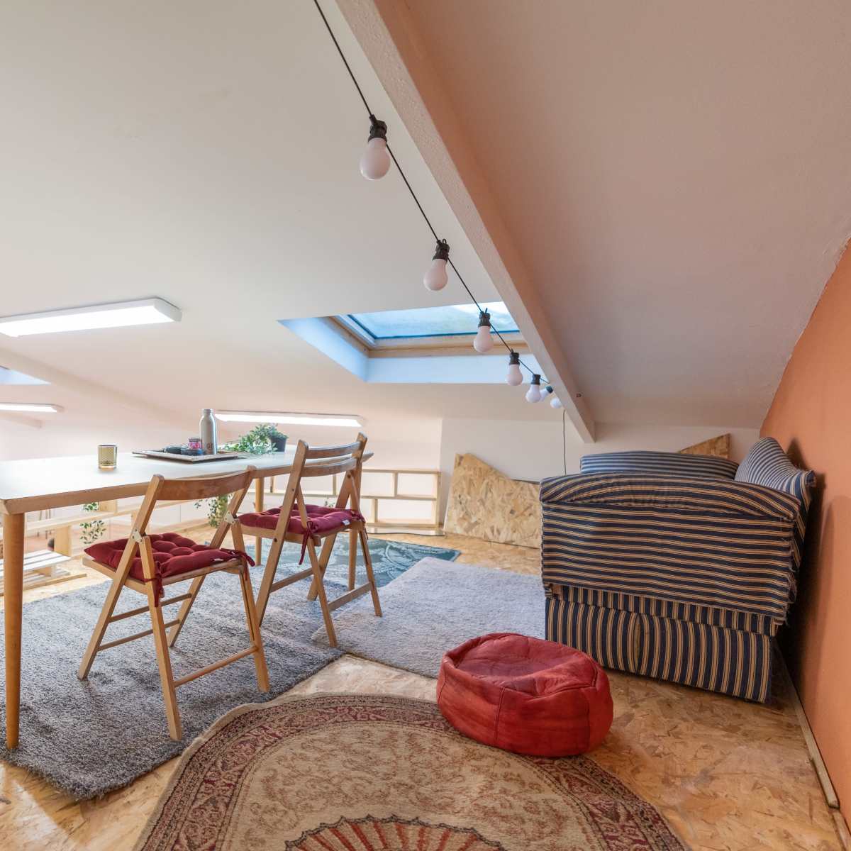 A cozy loft space featuring a wooden table with four chairs, a striped sofa, and potted plants under a skylight.