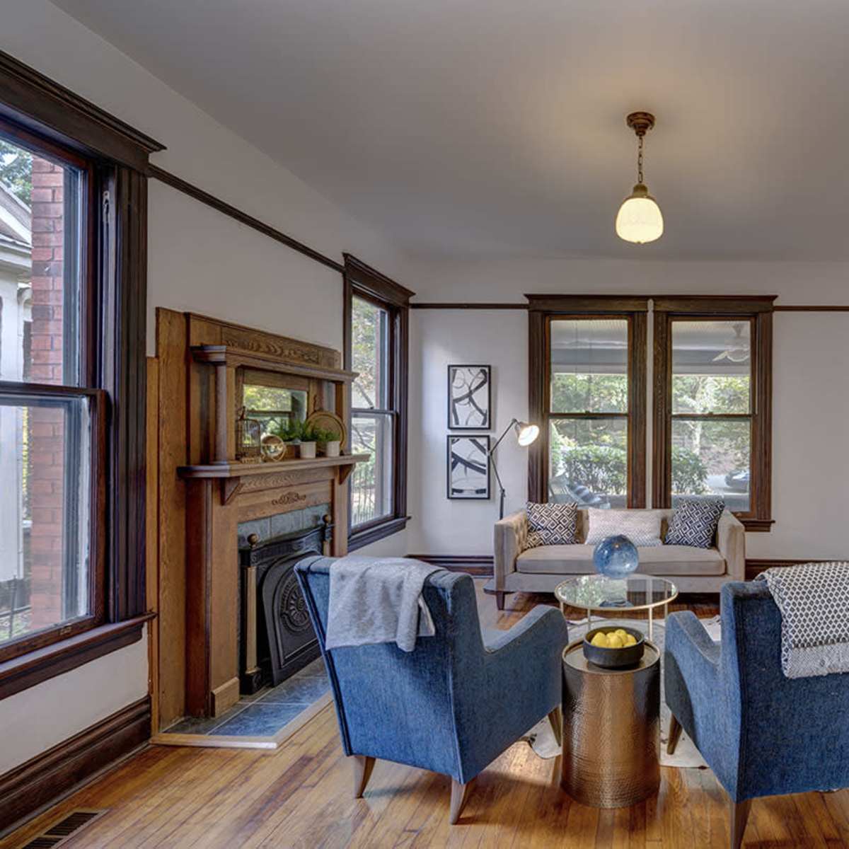 A cozy living room features two blue armchairs facing a round coffee table, with large windows allowing light to fill the space and a fireplace along one wall.