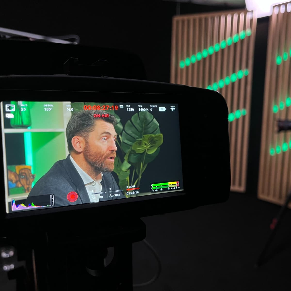 A camera displays a man speaking in a studio with decorative wooden panels and green lighting in the background.