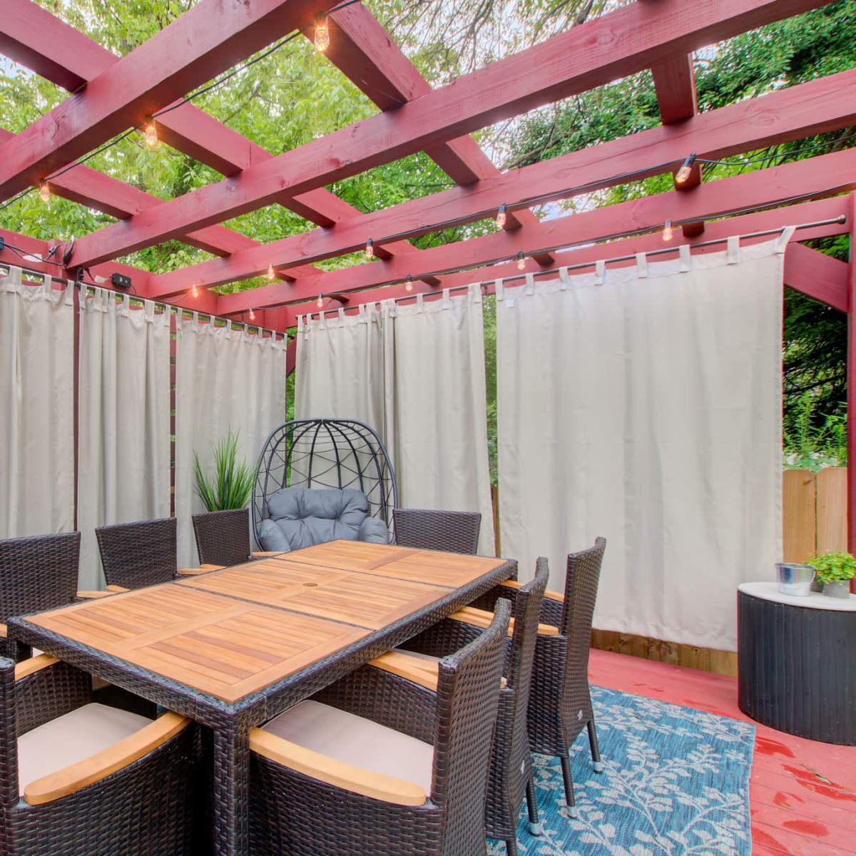 A shaded outdoor dining area with a wooden table surrounded by wicker chairs and hanging egg-shaped chairs, enclosed by light-colored curtains.
