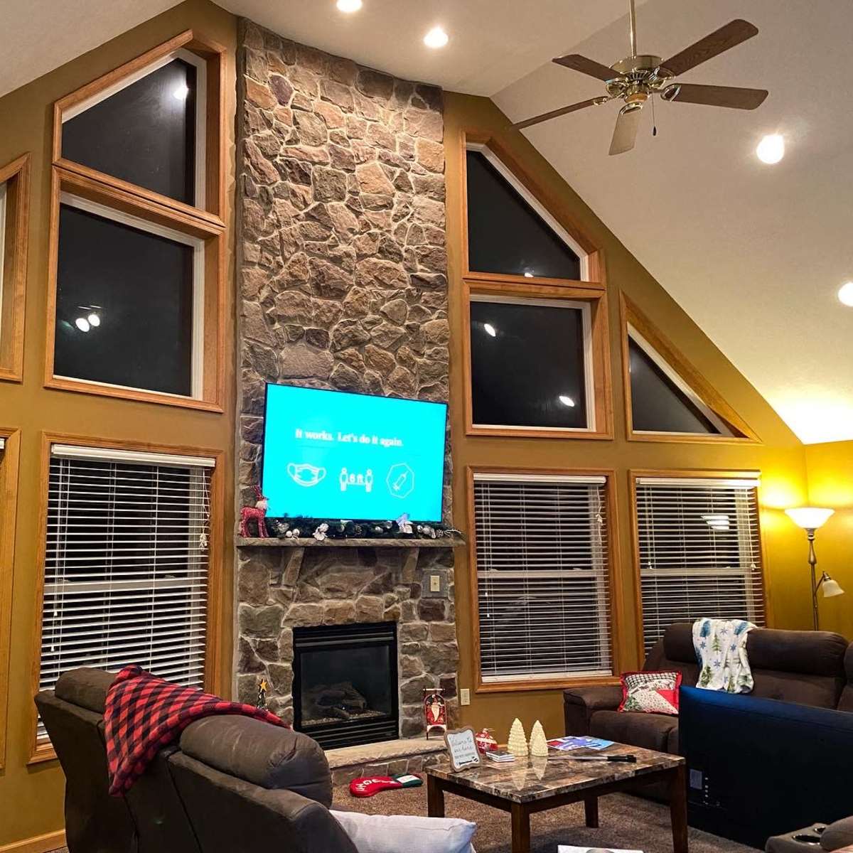 The scene shows a living room with a stone fireplace, large windows, a Christmas tree, and a mounted television displaying a message.