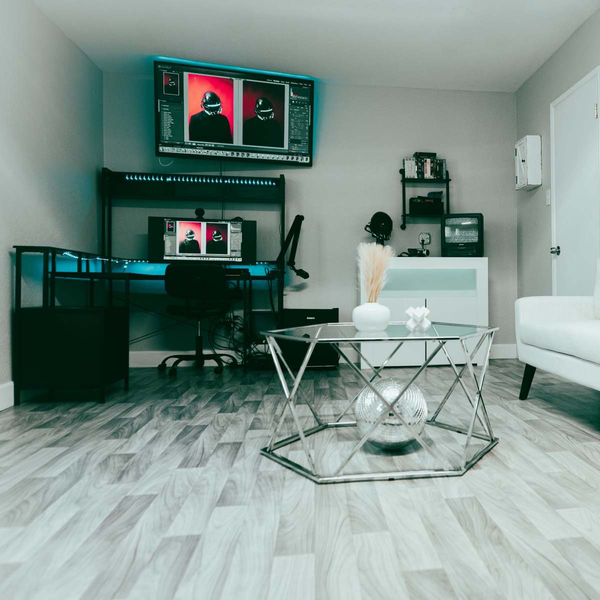 The room features a modern white sofa, a hexagonal glass coffee table, and a workspace setup with a computer and design tools against a light-colored wall.