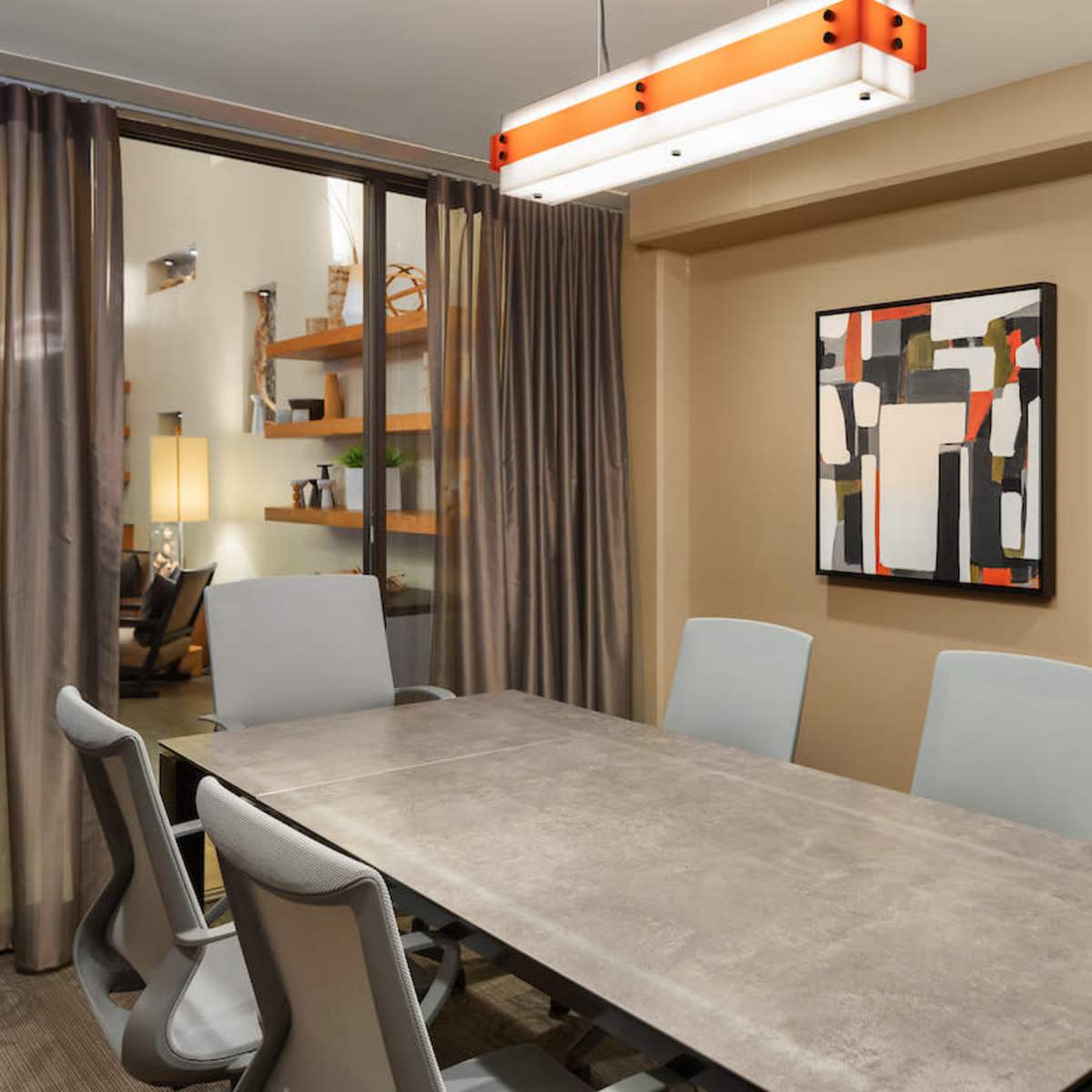 A modern conference room features a long table surrounded by light gray chairs, with two abstract paintings on the walls and a hanging light fixture overhead.