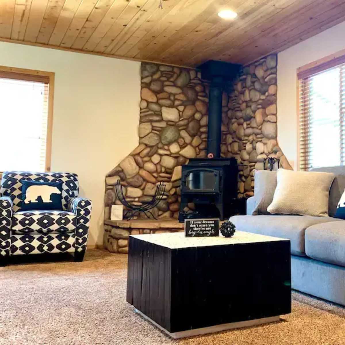 The living room features a gray sofa, a black and white patterned armchair, a wooden coffee table, and a stone fireplace with a wood stove.