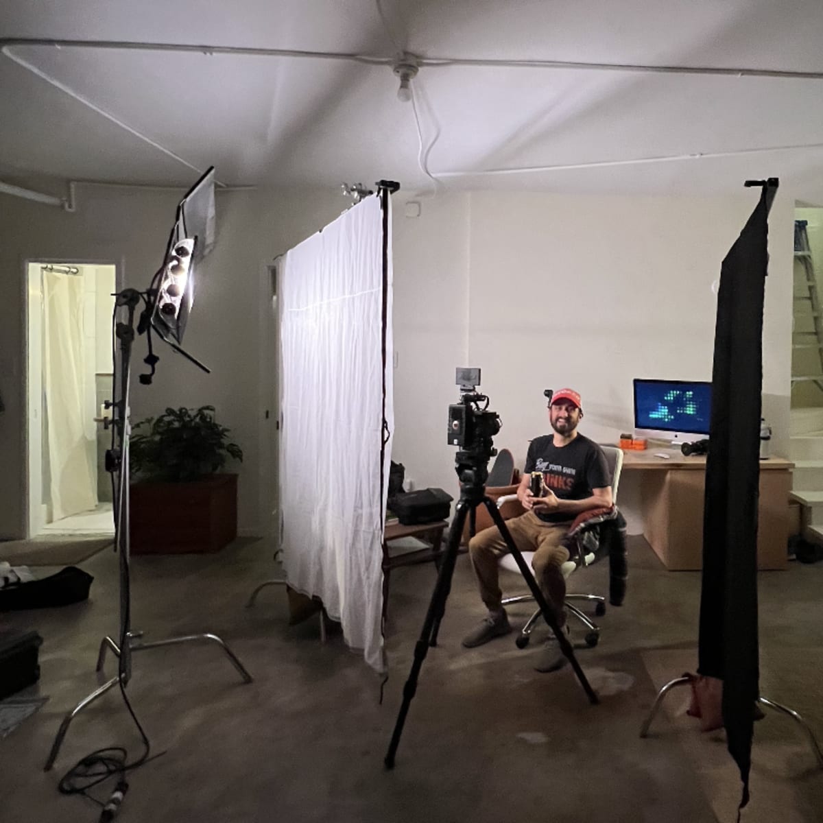 A person sits on a chair in a room set up for video recording, surrounded by lighting equipment and backdrops.
